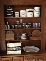 A wooden cabinet shelf holding 5 ceramic cookie jars with labels (Tea, Sugar, Flour, Cookies, and one unlabeled), various small cups like 4 ceramic handleless mugs, 4 farm scene mugs, 5 shallow goblets, 5 branded drinking glasses, two hurricane lamp tops, wooden salad serving set parts, and some boxes with chargers.
