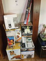 Photo showing stacked kitchen appliances including Keurig coffee maker, Oster rice cooker and bread maker in boxes, juicer box, Handy Steamer box, and storage organizer