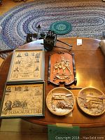 Overview of all items on wooden table with braided rug in background, showing two framed prints, carved wooden plaques, and metal cart