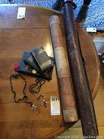View of worn leather fly fishing rod case, paper tube with German handwriting, 3 black bibles, and 2 rosaries all arranged on a wooden table.