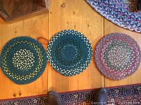 Three small circular braided rugs in blue, green, yellow, and purple color tones shown on wood floor.