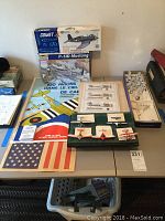 Photo of table displaying model airplane kits, mini planes, aviation posters including American flag and French aviation show, and airplane parts posters along with mounting shelf with mini planes.