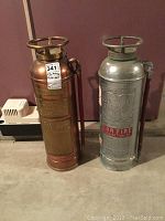 Photo of two antique fire extinguishers, one brass and one silver metal, showing overall appearance and size.