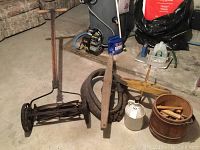 Overview of all items on concrete floor: manual reel mower, leather horse harness and yoke, wooden water bucket, pottery jug, and wagon wheel