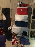 Stacked coolers: red Coleman cooler, blue cooler, large white foam cooler, with red serving bowls and metal canisters on top; folded grey EZ Up tent in black carry bag standing beside them; USA yard flags with wooden poles in front; plaid outdoor blanket partially visible on floor.