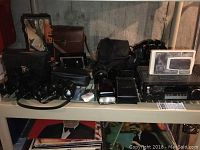 Wide shelf view showing the Minolta camera with lenses, original Polaroid camera with worn leather pouch, two binoculars (one Bushnell), camera flash, and two vintage radios