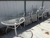 White round glass top metal patio table and eight white metal frame vinyl strap chairs stacked beside a white fence on outdoor deck, cushions not visible (stored separately).