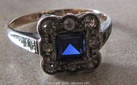 Front facing close-up showing square blue sapphire center with surrounding stones, some greyed and one missing, and yellow gold band sides.