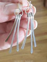 Close-up of the earrings held in a hand showing detail of the elongated flat silver bars attached to circular loops and pierced hooks.