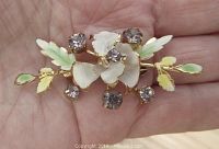 Top view of the brooch held in hand showing the gold tone flower design with prong set lavender rhinestones and hand painted enameled petals and leaves in white and green.