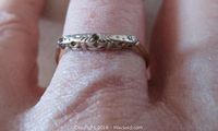 Close-up photo of the ring worn on a finger showing the engraved white gold top with one small diamond chip and two missing stones.