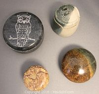 Four assorted containers shown from top view including a black soapstone box with owl carving, mottled green and brown round box, small handmade clay container and carved stone box.