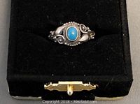 Photo of a sterling silver ring with ornate design and a small turquoise stone in the center, displayed on a black jewelry box.