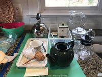 Showing the tea kettle, teapots, tea steeper, tea cups and saucers, wooden tea strainer and glass pitcher arranged on a kitchen counter