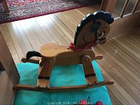 Child's wooden rocking horse with painted black mane, tail, large cartoon eyes, and red strap around neck shown from side angle on floor.