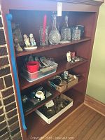 Wide view of wooden shelf displaying cut glass decanters, ceramic figurines, assorted glassware, and brass decor.