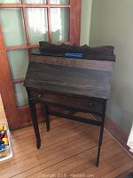 Full view of closed vintage secretary desk standing on a hardwood floor near a door; dark stained wood with visible wear and scratches.
