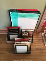 Wide shot showing a variety of picture frames stacked and wrapped in plastic on a wood floor against a wall.