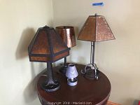 Four table lamps placed on a wooden round table in a corner, showing varied styles and sizes with metallic and hammered shades