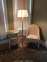 Full view of wicker chair, two-tiered wicker table, and turned-on floor lamp in a corner room with curtains and patterned carpet.