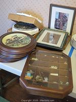 Various framed raccoon art, boxed plates, and stacked collectible plates on a table alongside a wooden glass-front wall curio filled with raccoon figurines