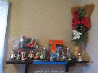 Wide shot of entire Christmas decor lot on wooden shelf including glass ornaments, miniature sleigh, Village Square Collection boxed hotel piece, and decorative broom with pine cones and red bow.