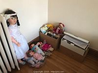 Wide view showing the large porcelain doll standing next to the wall and various smaller dolls and plush toys in cardboard boxes on wooden floor.