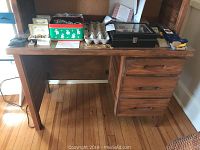 Wooden office desk with three drawers and a removable top hutch, placed against a wall on a wood floor. Various small items on the desk surface are excluded from sale.