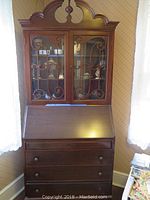 Front view of vintage secretary desk showing dark stained wood, glass display cabinet doors with decorative scrollwork, and three drawers below.
