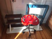 Red plastic saddle chair with holes in seat next to the shelving unit with three shelves of varied size and color.