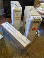 Two upright white Lakewood oil-filled space heaters on wheels and boxed Marvin window screen fan in room setting.