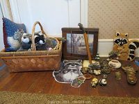 Full view of raccoon collection including basket with two stuffed raccoons, pillow, framed art, walking stick, wooden raccoon wall art, and multiple small raccoon-themed items arranged on floor