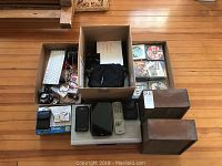 Full view of electronic items on wooden floor including boxed camera, Galaxy S6 phone, NEC projector, homemade speakers, Wii console, remote control, and CDs.