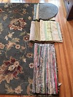 Photo showing one large floral area rug with multiple smaller rolled rugs including a heart-shaped one on hardwood floor
