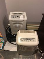 Two white dehumidifiers shown. Rear one is Maytag model M7DH45B2A, front one is GE model ADEL30LRQ2. Both have control panels on top front.