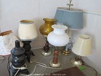 Seven assorted table lamps including blue fabric shade lamp, hobnail white milk glass shade lamp, amber glass lamp, and multiple lamps with cream or yellow fabric shades.