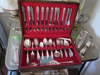 Silver plate flatware set arranged in a presentation box with red lining and additional cutlery in plastic sleeves and piles on the side