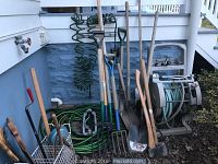 Full view of garden tools including hoses, shovels, pitchforks, axe, hose caddie and sprinklers stored outside.