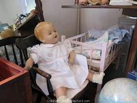 Vintage doll seated in child's rocking chair beside pink wooden toy crib with bedding.