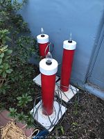 Three handmade outdoor electric candles displayed standing upright with red cylindrical bodies and white tops, with electrical cords visible, outdoors on dirt ground near plants.