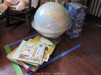 Photo showing large vintage globe on plastic base with pile of children's books underneath, including visible colorful covers and a large blue educational box to the right.