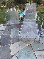 Three pieces of wrought iron outdoor furniture: one chaise lounge and two captain chairs placed on a stone patio, showcasing their full structure and design.