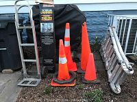 Photo showing hand truck, extension ladder stabilizer box, four orange traffic cones, and folded extension ladder against a blue wall outside.