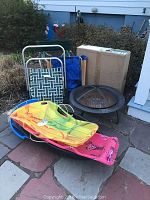Photo of the fire pit, snow sleds in various bright colors stacked together, three beach chairs, and a cardboard box of the fire pit nearby on stone pavement outside.