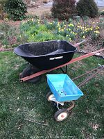 Side view of metal True Temper wheelbarrow and blue lawn spreader on grass near flower garden.