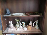 Photo showing various decorative vases, pitchers, two bells, a small trinket box, and two plates on wooden shelving.