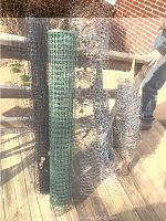 Five rolls of fencing material on wooden deck: 2 metal chicken wire, 1 green plastic fencing, 2 black plastic netting.