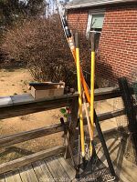 Photo showing the extendable pole tree limb lobber, rake, hoe, and other garden tools leaning against a wooden fence outdoors.