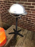 Full side view of the electric outdoor grill on a wooden deck against a brick wall, showing silver dome lid and black stand.