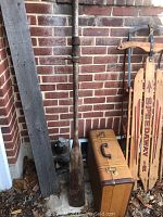 Photo showing the vintage Speedaway wooden runner sled with metal frame, rusted parts and broken runner board, standing next to a wooden oar and an old suitcase on the ground against a brick wall.